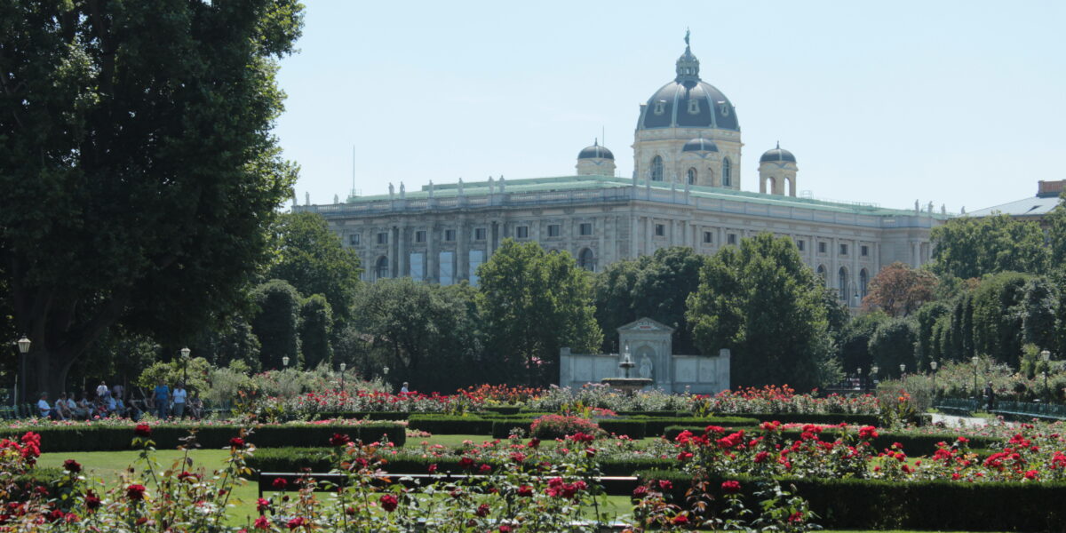 Ein großes historisches Gebäude mit Kuppel steht hinter einem formalen Garten mit roten Blumen und gestutzten Hecken, umgeben von Bäumen unter einem klaren Himmel.