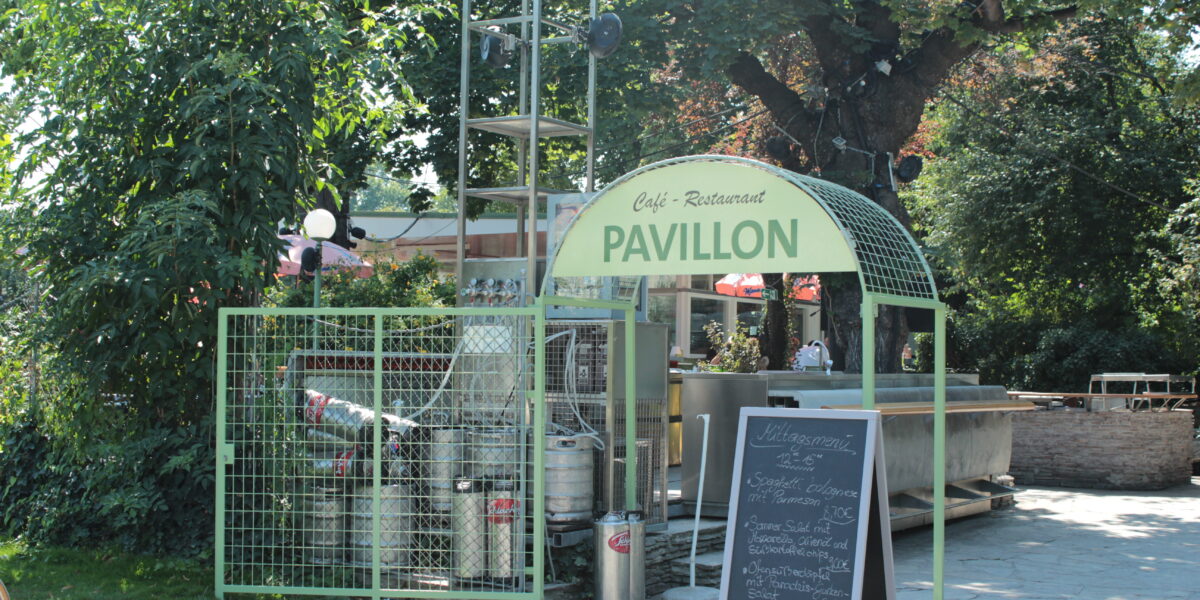 Außenansicht eines Café-Restaurants mit dem Namen Pavillon, mit Metallbierfässern, einer Speisekarte auf einer Kreidetafel und Bäumen im Hintergrund.