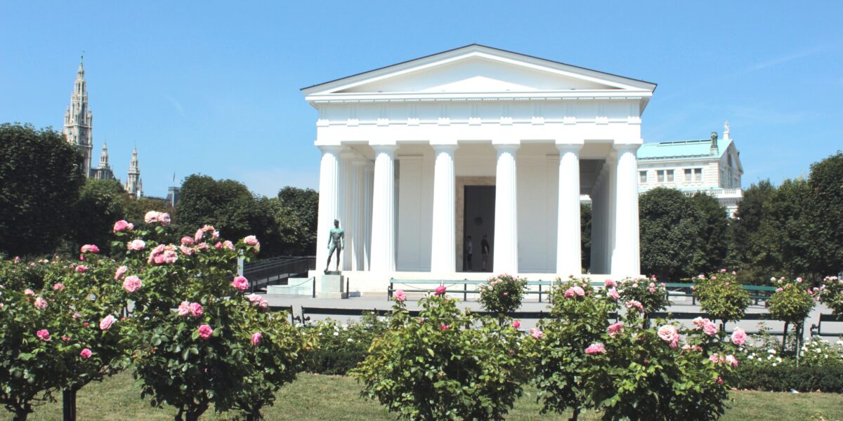 Ein weißes neoklassizistisches Gebäude im griechischen Stil mit Säulen steht in einem Garten mit blühenden rosa Rosen unter einem klaren blauen Himmel.