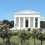 Ein weißes neoklassizistisches Gebäude im griechischen Stil mit Säulen steht in einem Garten mit blühenden rosa Rosen unter einem klaren blauen Himmel.