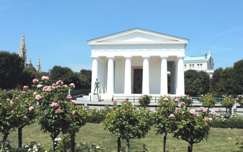 Ein weißes neoklassizistisches Gebäude im griechischen Stil mit Säulen steht in einem Garten mit blühenden rosa Rosen unter einem klaren blauen Himmel.