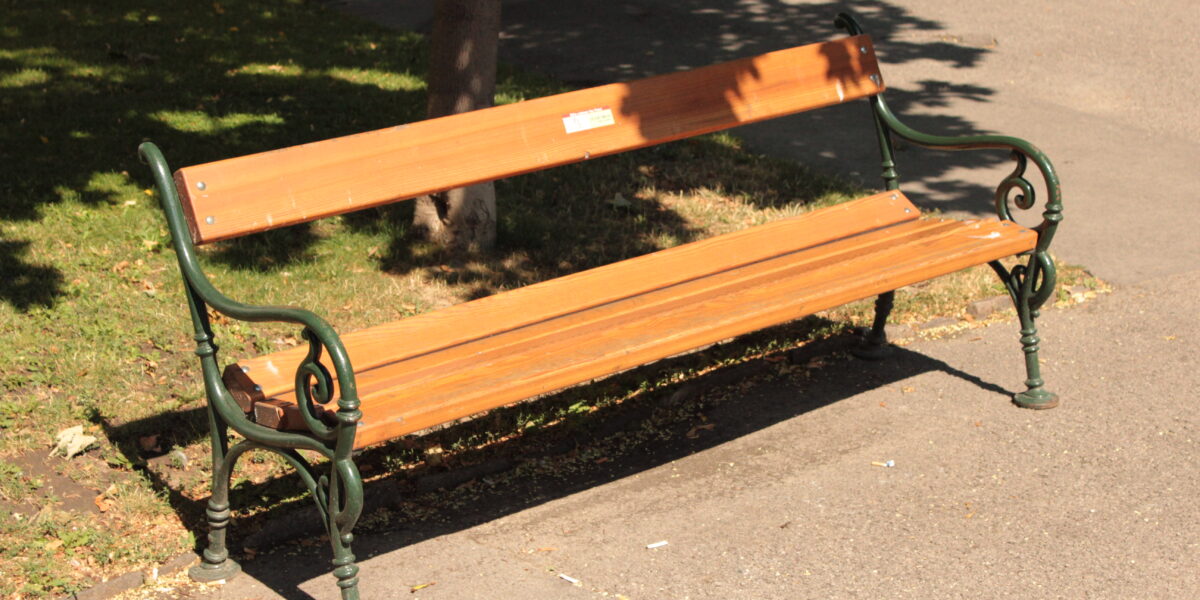 Eine hölzerne Parkbank mit Metallarmlehnen steht auf einem Gehweg neben Gras und einem Baum in hellem Sonnenlicht.