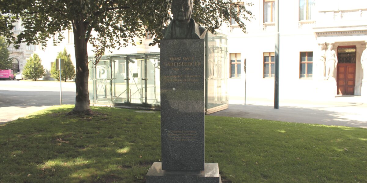 Eine Granitbüste steht auf einer Wiese in der Nähe eines Baumes, mit einem Gebäude und einer Glaskonstruktion im Hintergrund. Auf dem Sockel ist ein Text eingraviert.
