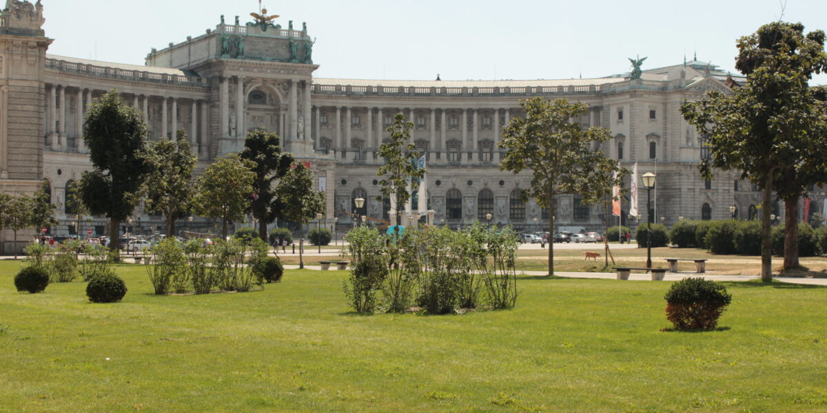 Neoklassizistisches Palastgebäude mit Statuen und Säulen, an einem sonnigen Tag von einem grasbewachsenen Park mit Bäumen und Sträuchern im Vordergrund aus gesehen.