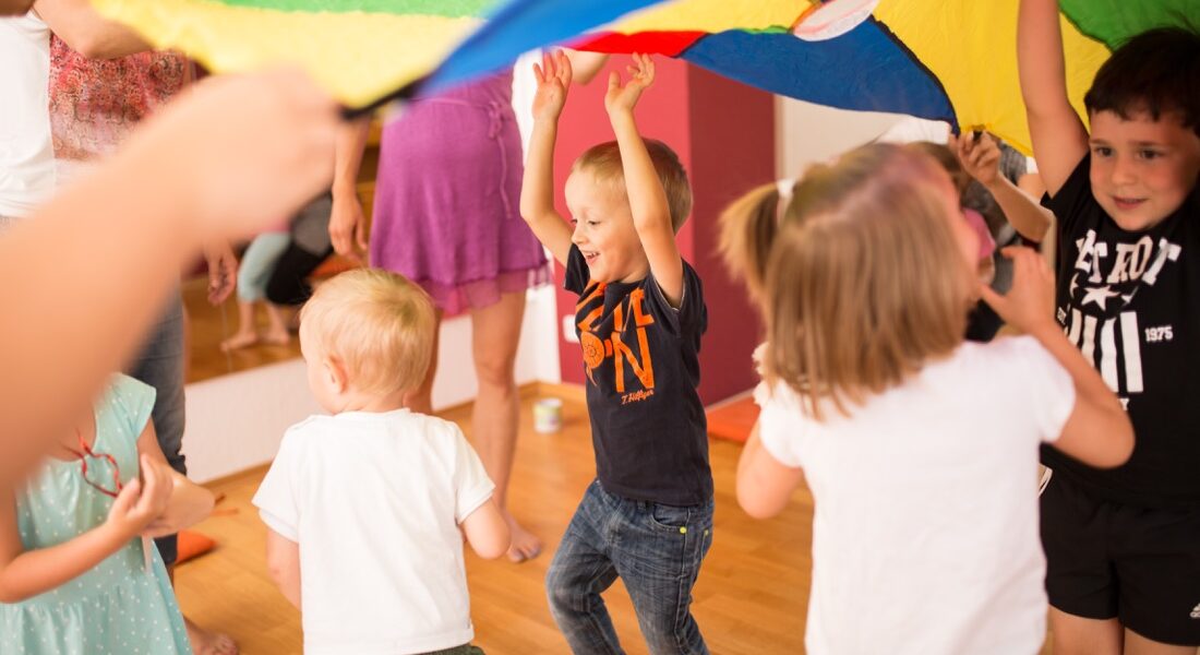 Kinder spielen drinnen zusammen, halten einen bunten Fallschirm hoch und bewegen ihn. Einige Erwachsene sind anwesend und helfen den Kindern. Der Raum hat Holzböden und helle Wände.