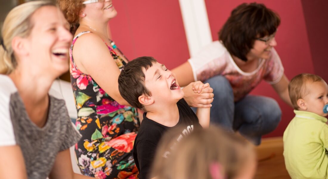 Eine Gruppe von Erwachsenen und Kindern sitzt oder hockt drinnen und lacht und lächelt gemeinsam vor einer roten Wand.