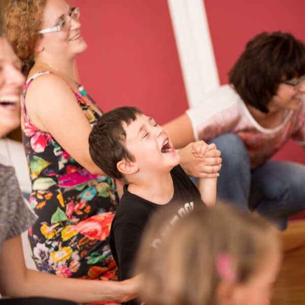 Eine Gruppe von Erwachsenen und Kindern sitzt oder hockt drinnen und lacht und lächelt gemeinsam vor einer roten Wand.