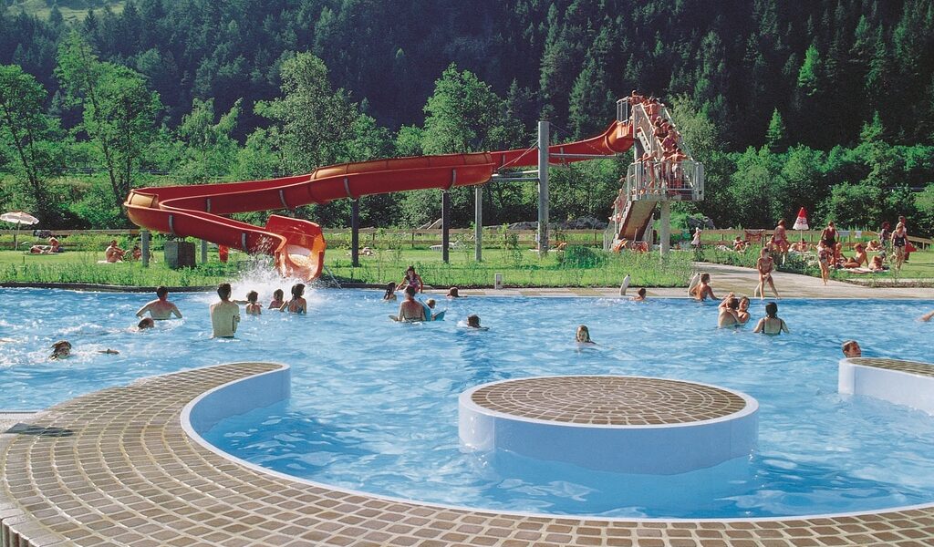 Die Menschen schwimmen und entspannen in einem Außenpool mit einer roten Wasserrutsche, umgeben von grünen Bäumen und Bergen im Hintergrund.