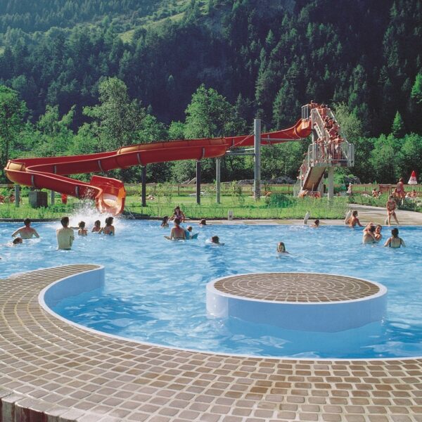 Die Menschen schwimmen und entspannen in einem Außenpool mit einer roten Wasserrutsche, umgeben von grünen Bäumen und Bergen im Hintergrund.
