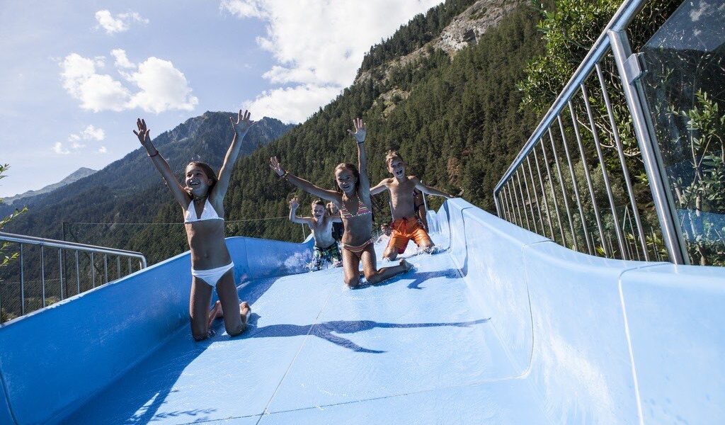 Vier Kinder rutschen im Freien, umgeben von Bäumen und Bergen unter einem teilweise bewölkten Himmel, eine blaue Wasserrutsche hinunter.