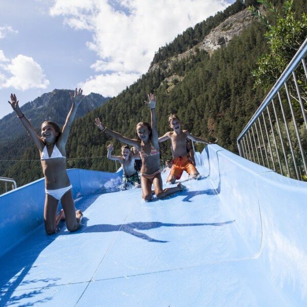 Vier Kinder rutschen im Freien, umgeben von Bäumen und Bergen unter einem teilweise bewölkten Himmel, eine blaue Wasserrutsche hinunter.