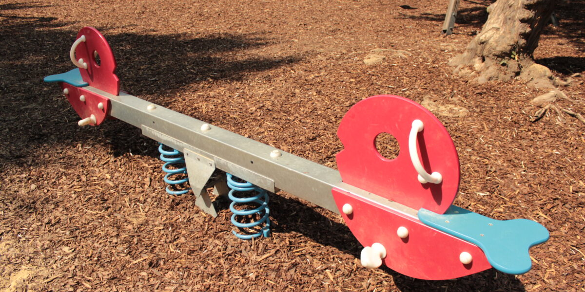Eine rote und blaue gefederte Wippe steht auf Holzspänen auf einem Spielplatz, während im Hintergrund weitere Spielgeräte zu sehen sind.