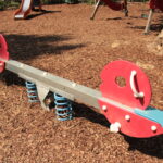 Eine rote und blaue gefederte Wippe steht auf Holzspänen auf einem Spielplatz, während im Hintergrund weitere Spielgeräte zu sehen sind.