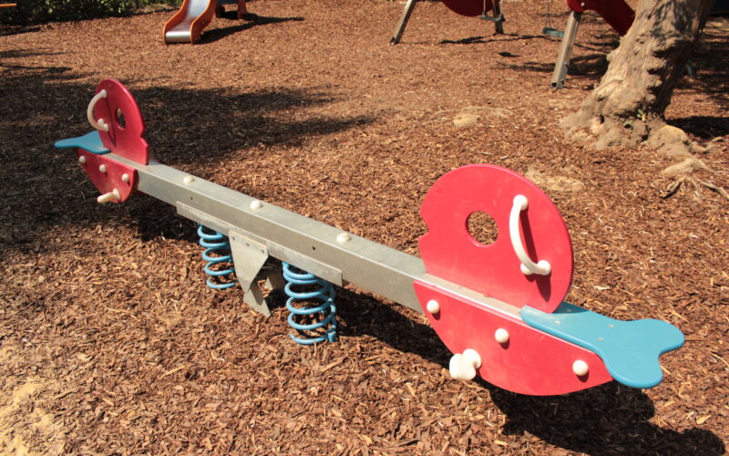 Eine rote und blaue gefederte Wippe steht auf Holzspänen auf einem Spielplatz, während im Hintergrund weitere Spielgeräte zu sehen sind.