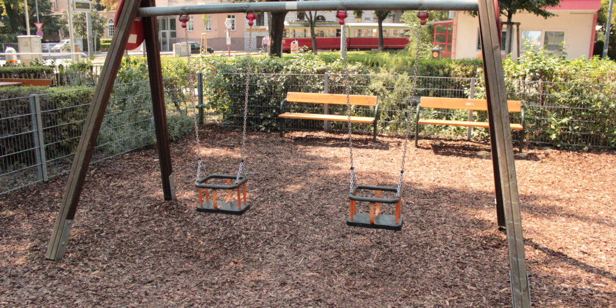 Zwei leere Schaukeln für Kleinkinder hängen an einem Holzrahmen auf einem Spielplatz, mit Bänken, Zäunen und Grünpflanzen im Hintergrund.