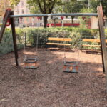Zwei leere Schaukeln für Kleinkinder hängen an einem Holzrahmen auf einem Spielplatz, mit Bänken, Zäunen und Grünpflanzen im Hintergrund.