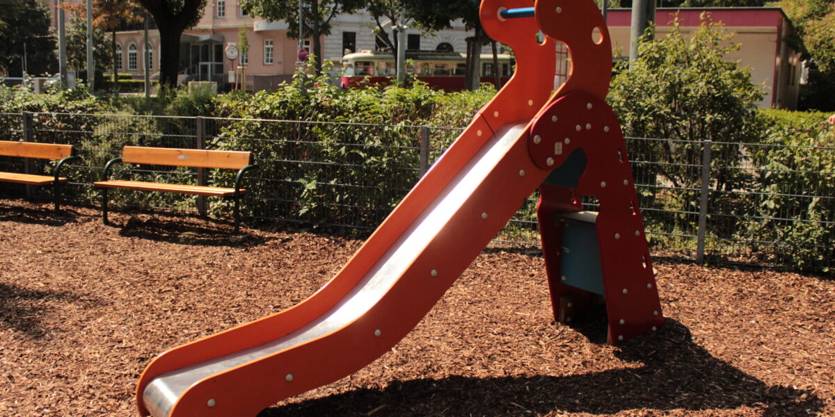 Eine kleine rote Kinderrutsche mit einer Metalloberfläche steht auf Holzspänen auf einem Spielplatz, mit Bänken und Grünpflanzen im Hintergrund.
