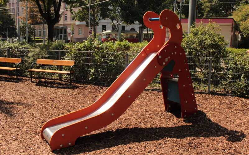Eine kleine rote Kinderrutsche mit einer Metalloberfläche steht auf Holzspänen auf einem Spielplatz, mit Bänken und Grünpflanzen im Hintergrund.