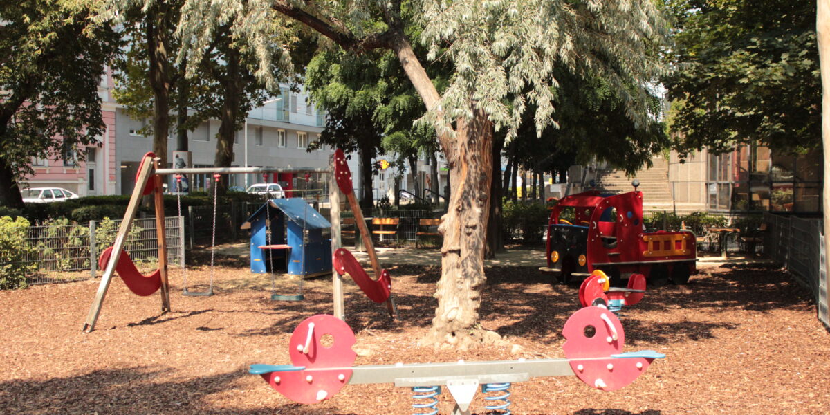 Ein Spielplatz mit einer Wippe, Schaukeln, einer Rutsche und einem roten Spielzeug-Feuerwehrauto, umgeben von Bäumen und Holzspänen.