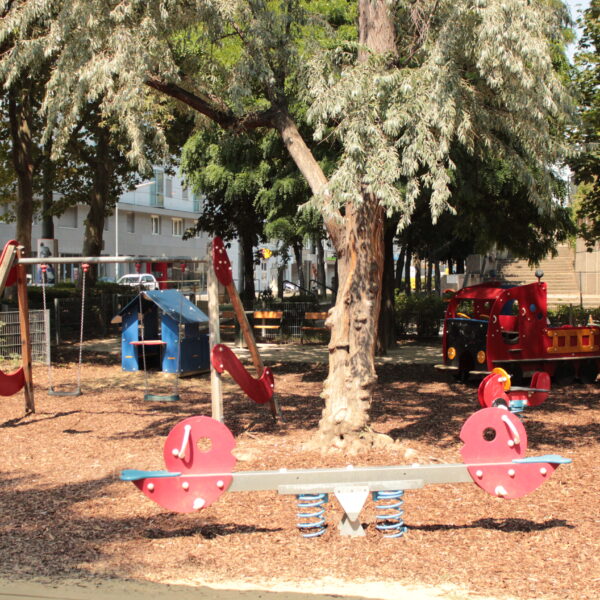 Ein Spielplatz mit einer Wippe, Schaukeln, einer Rutsche und einem roten Spielzeug-Feuerwehrauto, umgeben von Bäumen und Holzspänen.