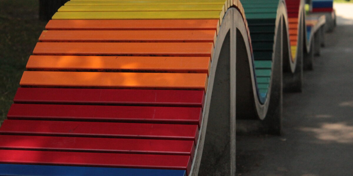 Eine wellenförmige, regenbogenfarbene Bank aus Beton und Holzlatten in einer Parkanlage.