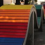 Eine wellenförmige, regenbogenfarbene Bank aus Beton und Holzlatten in einer Parkanlage.