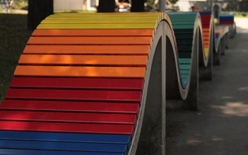 Eine wellenförmige, regenbogenfarbene Bank aus Beton und Holzlatten in einer Parkanlage.