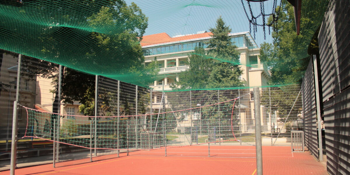 Ein roter Sportplatz mit Basketballkorb, Badmintonnetz und grünem Sicherheitsnetz, umgeben von einem Metallzaun und Bäumen.
