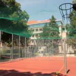 Ein roter Sportplatz mit Basketballkorb, Badmintonnetz und grünem Sicherheitsnetz, umgeben von einem Metallzaun und Bäumen.