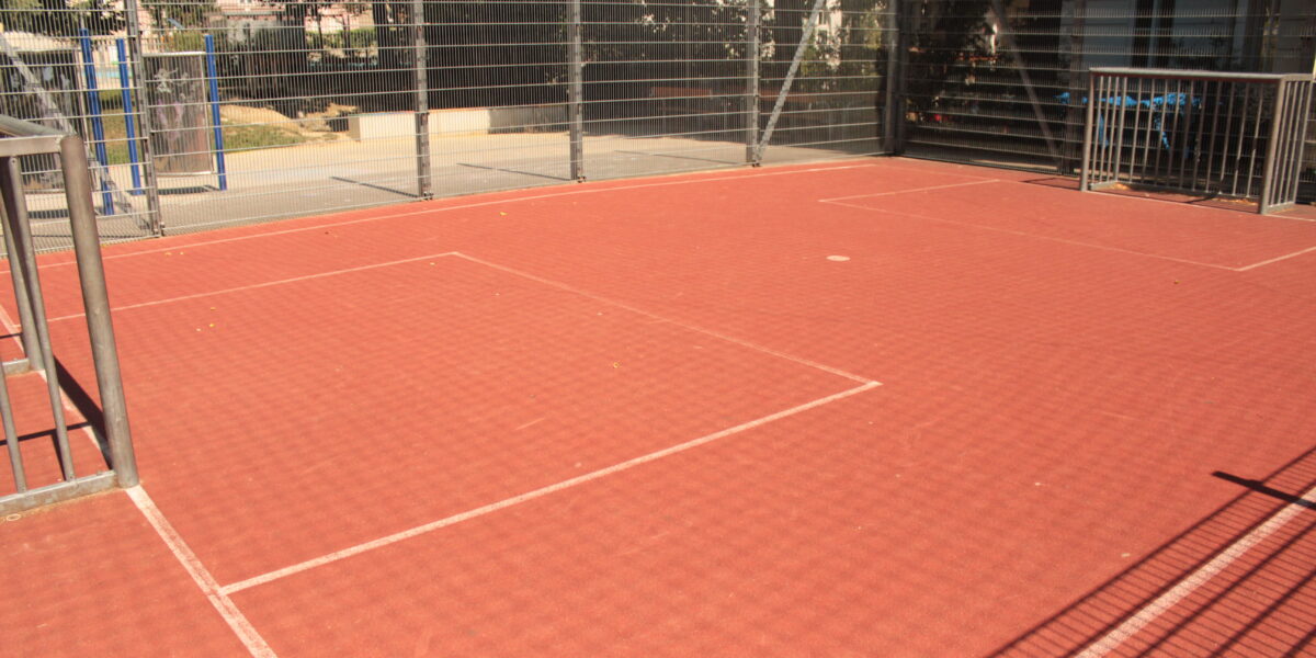 Ein eingezäunter Sportplatz im Freien mit rotem Belag und weißen Linien, umgeben von Metallgeländern und Maschendrahtzaun, im Sonnenlicht.