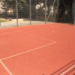 Ein eingezäunter Sportplatz im Freien mit rotem Belag und weißen Linien, umgeben von Metallgeländern und Maschendrahtzaun, im Sonnenlicht.