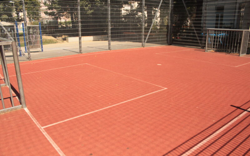 Ein eingezäunter Sportplatz im Freien mit rotem Belag und weißen Linien, umgeben von Metallgeländern und Maschendrahtzaun, im Sonnenlicht.