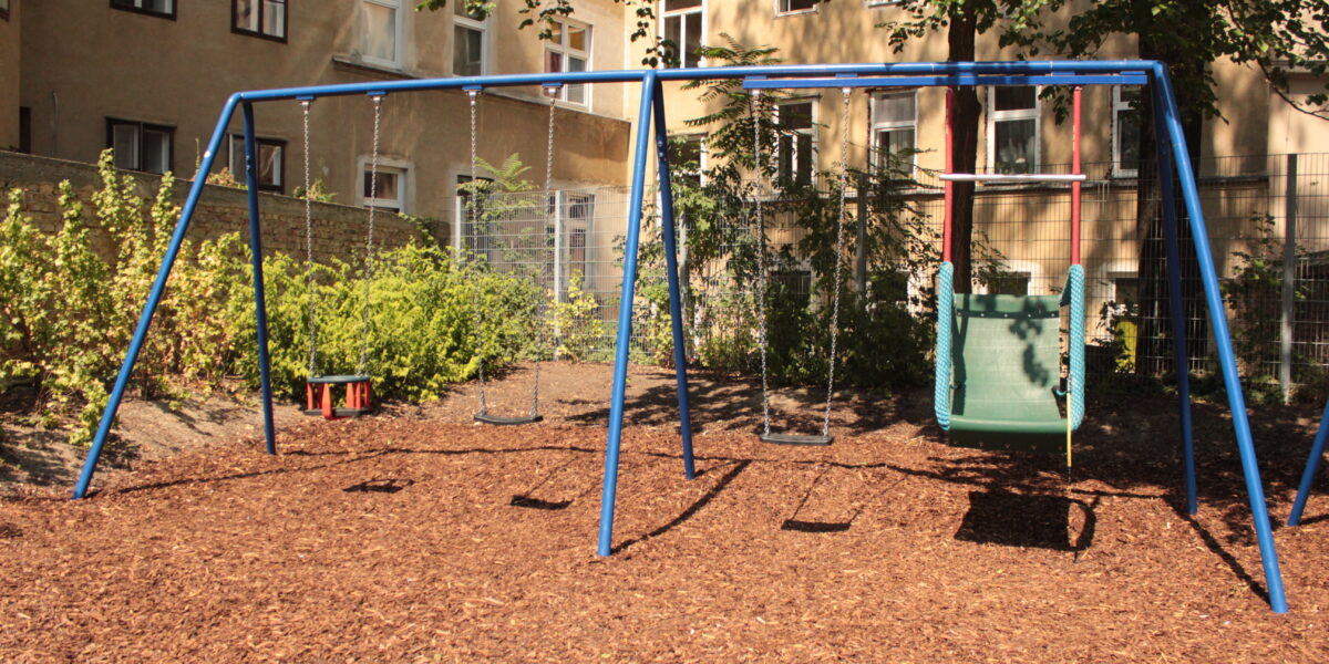 Ein Spielplatz mit blauen Metallschaukeln, darunter ein grüner barrierefreier Schaukelsitz, steht auf einer Mulchfläche in der Nähe von Sträuchern und einem alten Wohnhaus.