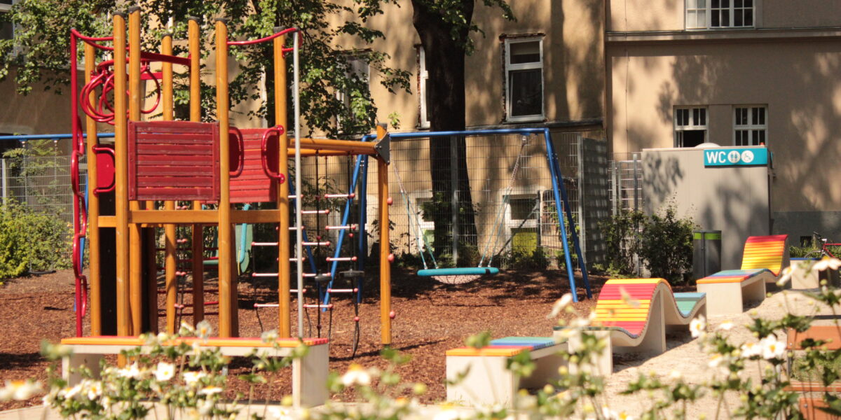 Ein Spielplatz mit einem Klettergerüst, Schaukeln und bunten Bänken inmitten von Grün; im Hintergrund sind ein Gebäude und ein Schild für Toiletten zu sehen.