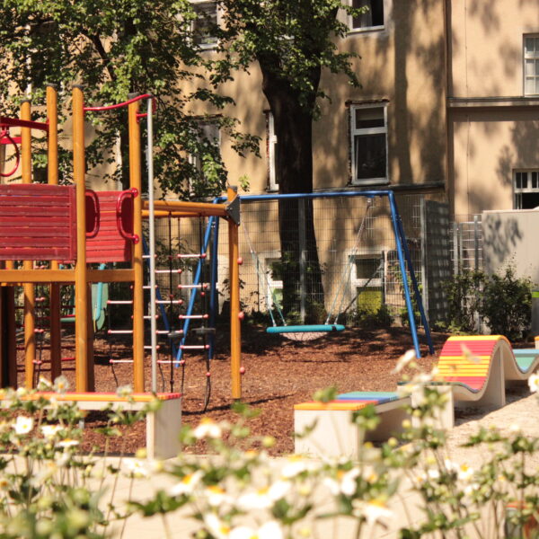 Ein Spielplatz mit einem Klettergerüst, Schaukeln und bunten Bänken inmitten von Grün; im Hintergrund sind ein Gebäude und ein Schild für Toiletten zu sehen.