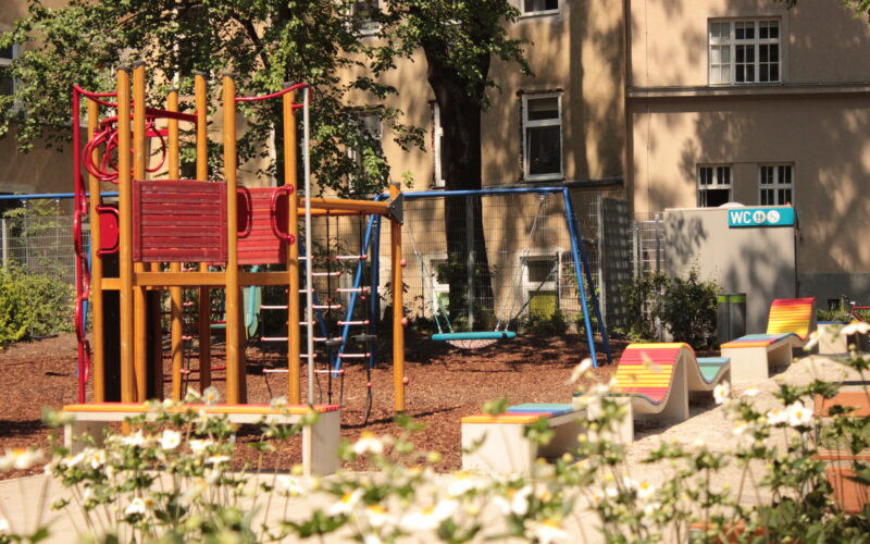 Ein Spielplatz mit einem Klettergerüst, Schaukeln und bunten Bänken inmitten von Grün; im Hintergrund sind ein Gebäude und ein Schild für Toiletten zu sehen.