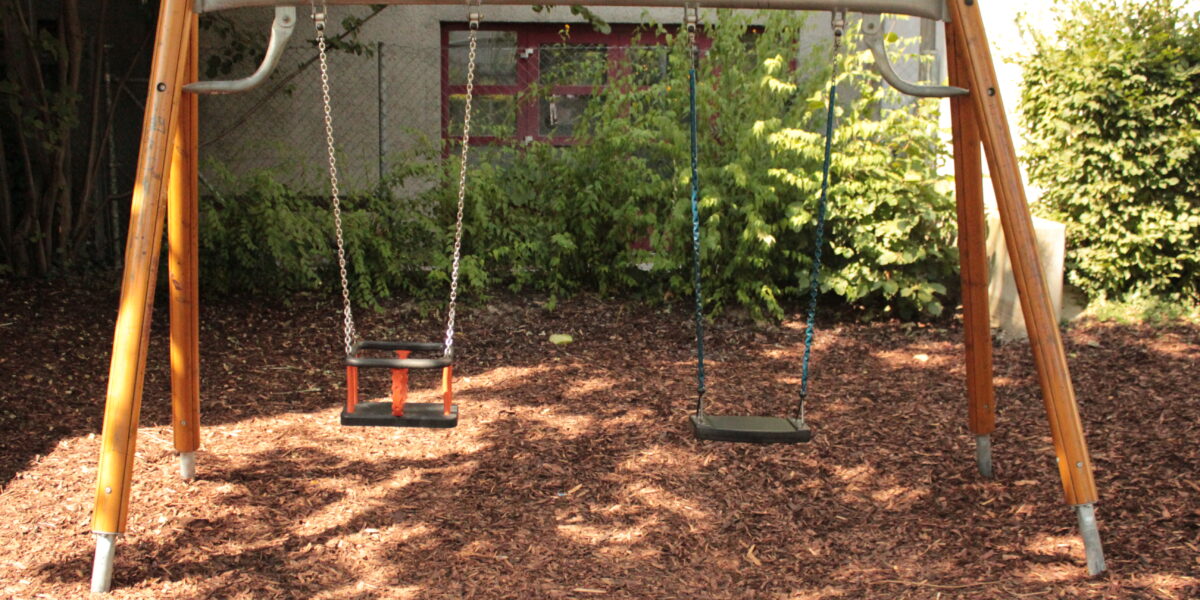 Eine Schaukel aus Metall und Holz mit zwei leeren Schaukeln steht auf einem mit Holzspänen bedeckten Spielplatz, mit Grünpflanzen und einem Gebäude im Hintergrund.