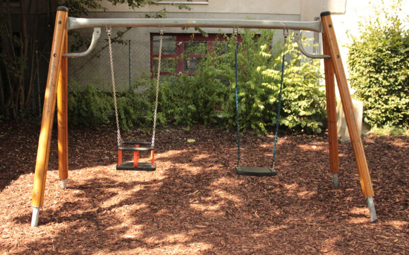 Eine Schaukel aus Metall und Holz mit zwei leeren Schaukeln steht auf einem mit Holzspänen bedeckten Spielplatz, mit Grünpflanzen und einem Gebäude im Hintergrund.