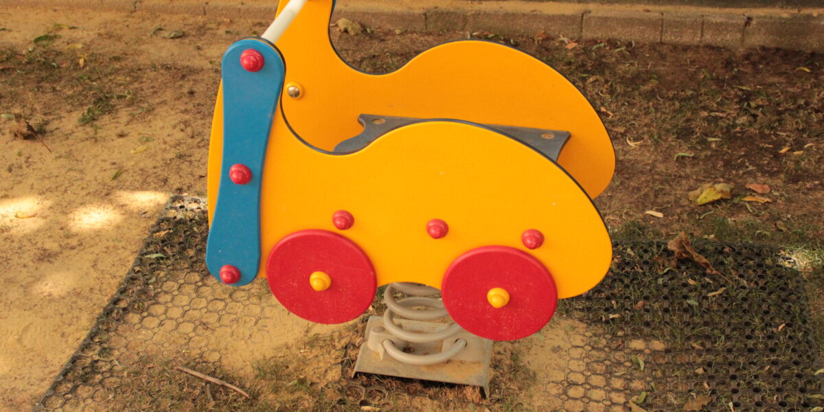 Ein gelber und roter Springreiter in Form eines Autos ist auf dem Sandboden eines Spielplatzes installiert.