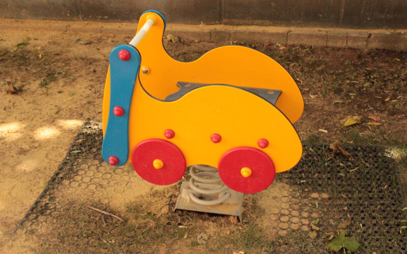 Ein gelber und roter Springreiter in Form eines Autos ist auf dem Sandboden eines Spielplatzes installiert.