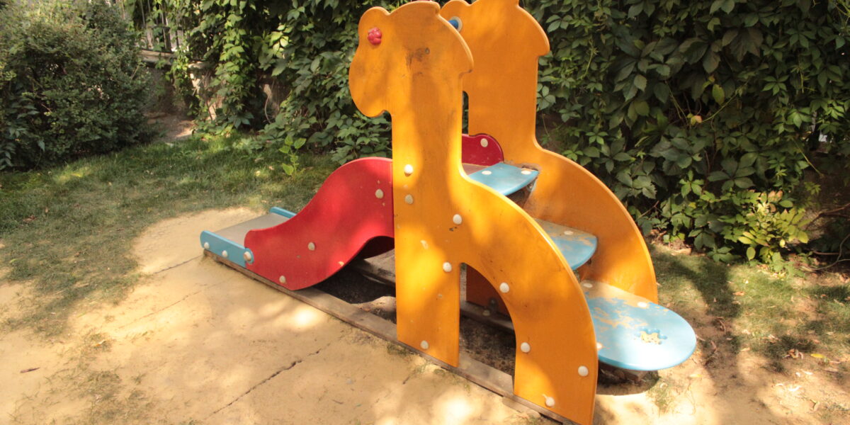 Eine kleine Spielplatzrutsche mit gelben Seiten in Form von Giraffen, blauen Stufen und einer roten Rutschfläche, die auf einem Stück Sand steht und von grünen Büschen umgeben ist.