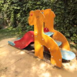 Eine kleine Spielplatzrutsche mit gelben Seiten in Form von Giraffen, blauen Stufen und einer roten Rutschfläche, die auf einem Stück Sand steht und von grünen Büschen umgeben ist.