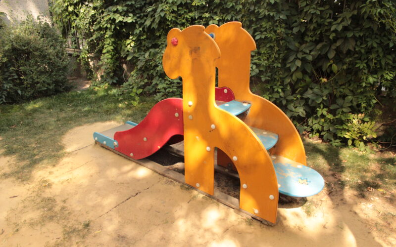 Eine kleine Spielplatzrutsche mit gelben Seiten in Form von Giraffen, blauen Stufen und einer roten Rutschfläche, die auf einem Stück Sand steht und von grünen Büschen umgeben ist.