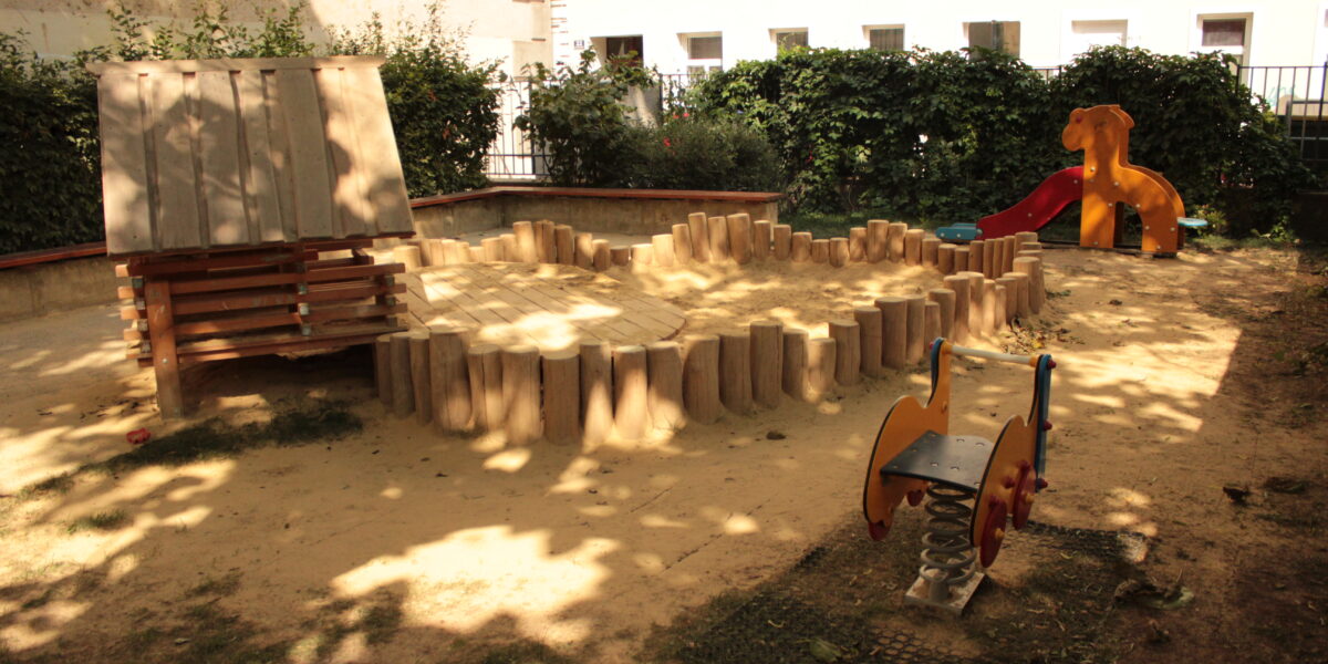 Ein kleiner Spielplatz mit einem Sandbereich, einem Holzspielgerät, einer orangefarbenen Rutsche und einem Springreiter, umgeben von Büschen und einem Gebäude im Hintergrund.