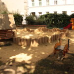 Ein kleiner Spielplatz mit einem Sandbereich, einem Holzspielgerät, einer orangefarbenen Rutsche und einem Springreiter, umgeben von Büschen und einem Gebäude im Hintergrund.