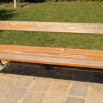 Eine abgenutzte Holzbank mit grünen Metallarmlehnen und -beinen steht auf einem Steinpflaster vor einer Wiese.