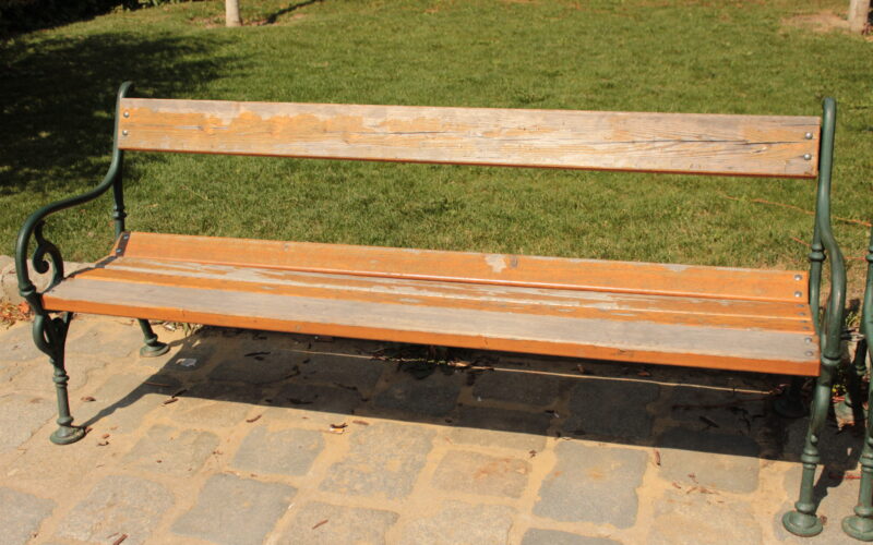 Eine abgenutzte Holzbank mit grünen Metallarmlehnen und -beinen steht auf einem Steinpflaster vor einer Wiese.