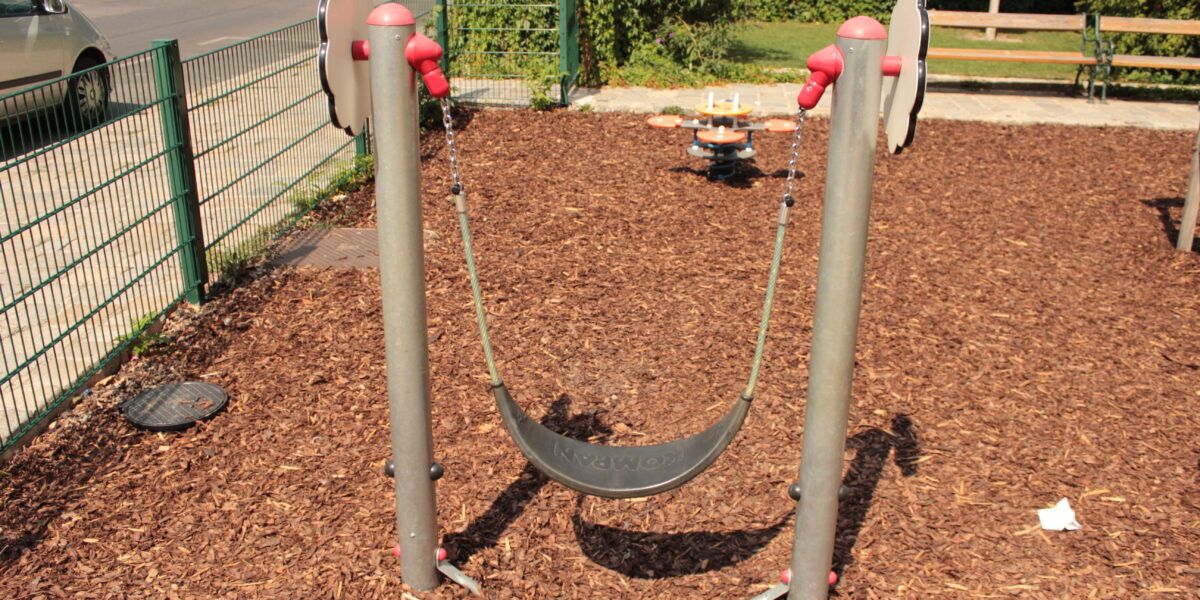 Ein flexibler Spielplatz-Schaukelsitz ist zwischen zwei Metallpfosten über einem Holzschnitzel-Bodenbelag aufgehängt; im Hintergrund sind ein Zaun und Bänke zu sehen.