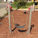 Ein flexibler Spielplatz-Schaukelsitz ist zwischen zwei Metallpfosten über einem Holzschnitzel-Bodenbelag aufgehängt; im Hintergrund sind ein Zaun und Bänke zu sehen.