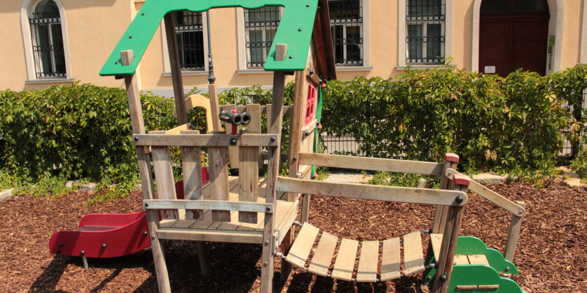Spielplatz aus Holz mit einer Rutsche, einer Kletterbrücke und einem Aussichtsbereich, der auf Mulch liegt, mit einer Hecke und einem Gebäude im Hintergrund.